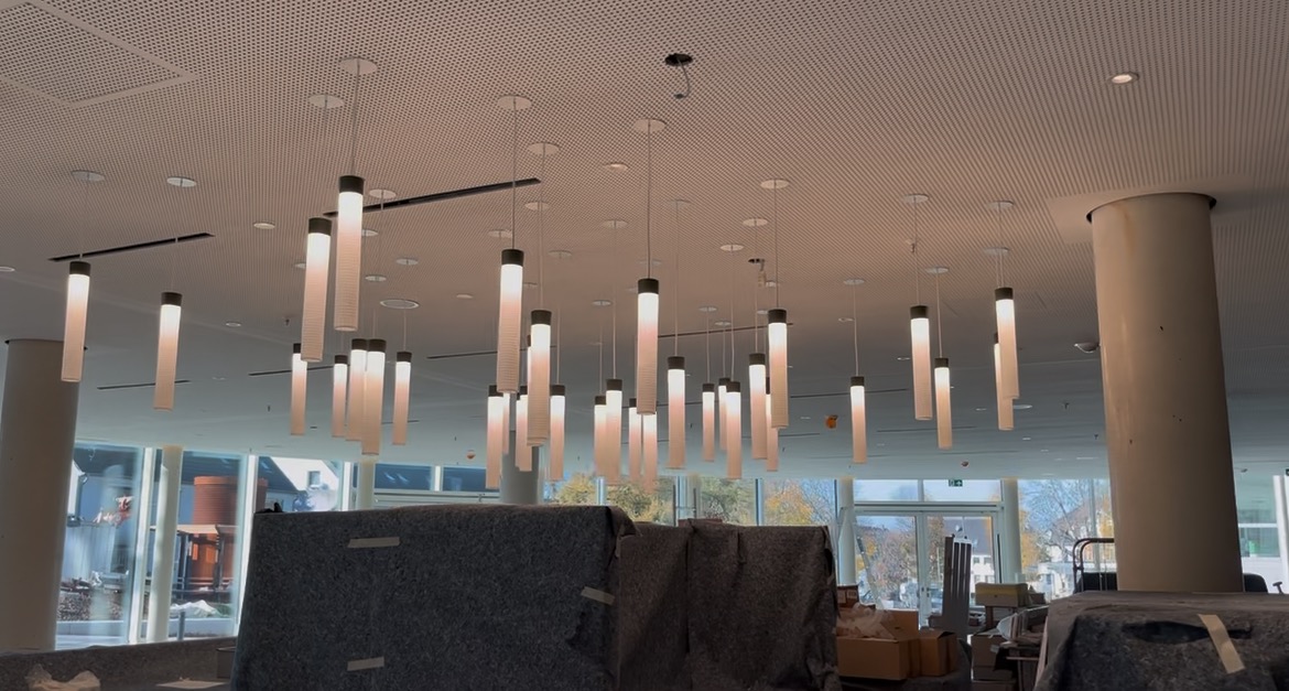 Nowoczesne oświetlenie sufitowe z cylindrycznymi lampami wiszącymi w jasnym wnętrzu, widoczne kolumny i okna z widokiem na zewnątrz. Częściowo zakryte meble.