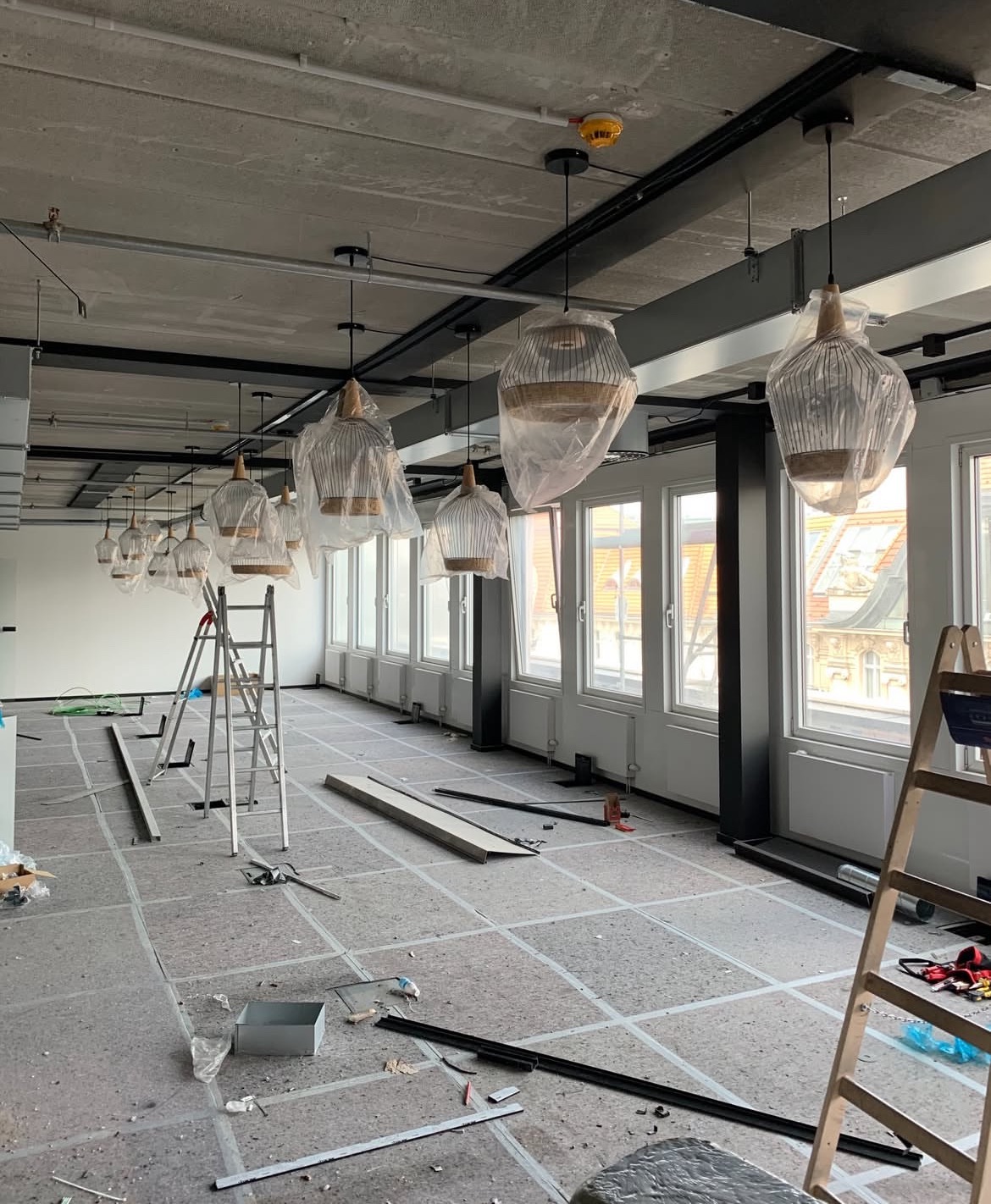 Wnętrze biura w trakcie wykończenia: podłoga zabezpieczona, wiszące lampy w folii ochronnej, drabiny, okna z widokiem na miasto. Etap prac wykończeniowych.