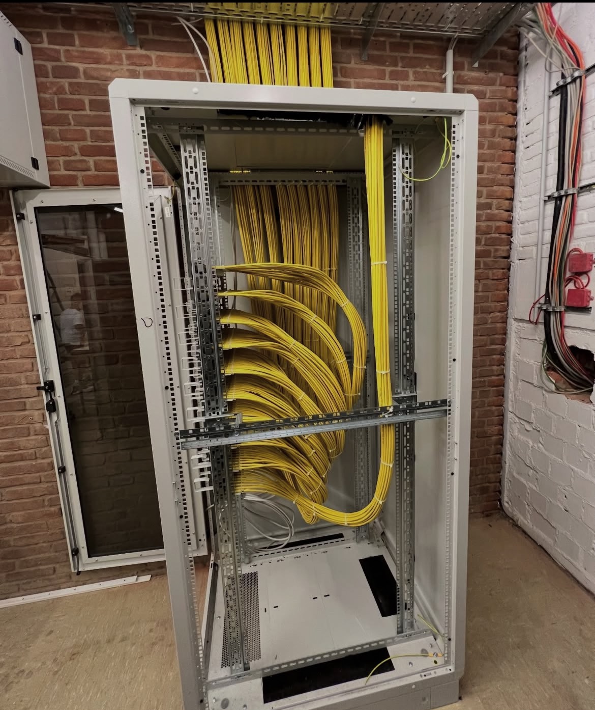 Szafa serwerowa z uporządkowanymi, żółtymi kablami. Widoczne elementy montażowe i fragment ściany z cegły. Estetyczne ułożenie przewodów wewnątrz szafy.
