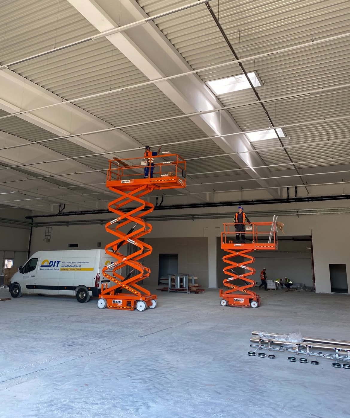 Montaż instalacji w hali: Dwóch pracowników na podnośnikach nożycowych, montuje elementy pod sufitem. Biała hala z widocznymi instalacjami i oświetleniem.