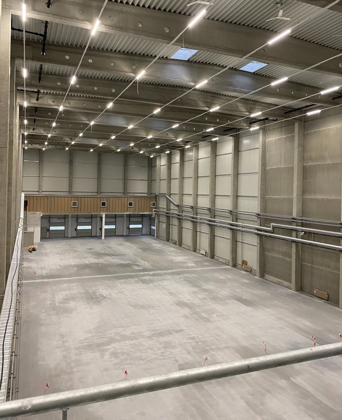 Przestronne wnętrze hali magazynowej z betonową podłogą, oświetleniem LED pod sufitem i bramami w tle. Widok z antresoli.