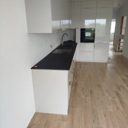 BUDREM OLHA MOROZ - Nowoczesna kuchnia w trakcie wykańczania: białe szafki z czarnym blatem, drewnopodobne płytki na podłodze, widoczne niedokończone ściany i drabina w tle.