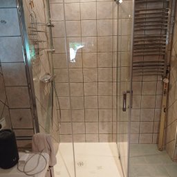 BUDREM OLHA MOROZ - Kabina prysznicowa z chromowanymi profilami i grzejnikiem drabinkowym na ścianie z beżowych płytek. Widoczny fragment umywalki i akcesoria łazienkowe.