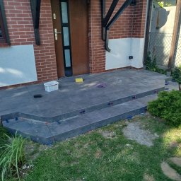 BUDREM OLHA MOROZ - Układanie ciemnych płytek na schodach wejściowych do domu. Widoczne narzędzia i elementy mocujące. Prace w toku, otoczenie zielone, trawa.