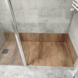 BUDREM OLHA MOROZ - Wykończona kabina prysznicowa z odpływem liniowym i płytkami imitującymi drewno na podłodze. Na ścianach szare płytki. Widoczny grzejnik drabinkowy.