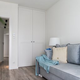 Wymarzone Meble - Biała szafa wnękowa z dwoma drzwiami, widoczna z boku szara sofa z poduszkami i pledem. W tle otwarte drzwi do kolejnego pomieszczenia. Minimalistyczny wystrój.