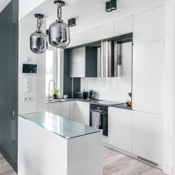 Wymarzone Meble - Nowoczesna, minimalistyczna kuchnia w bieli i szarości z wyspą z blatem ze szkła. Chromowane lampy wiszące i okap. Jasne drewniane panele podłogowe.