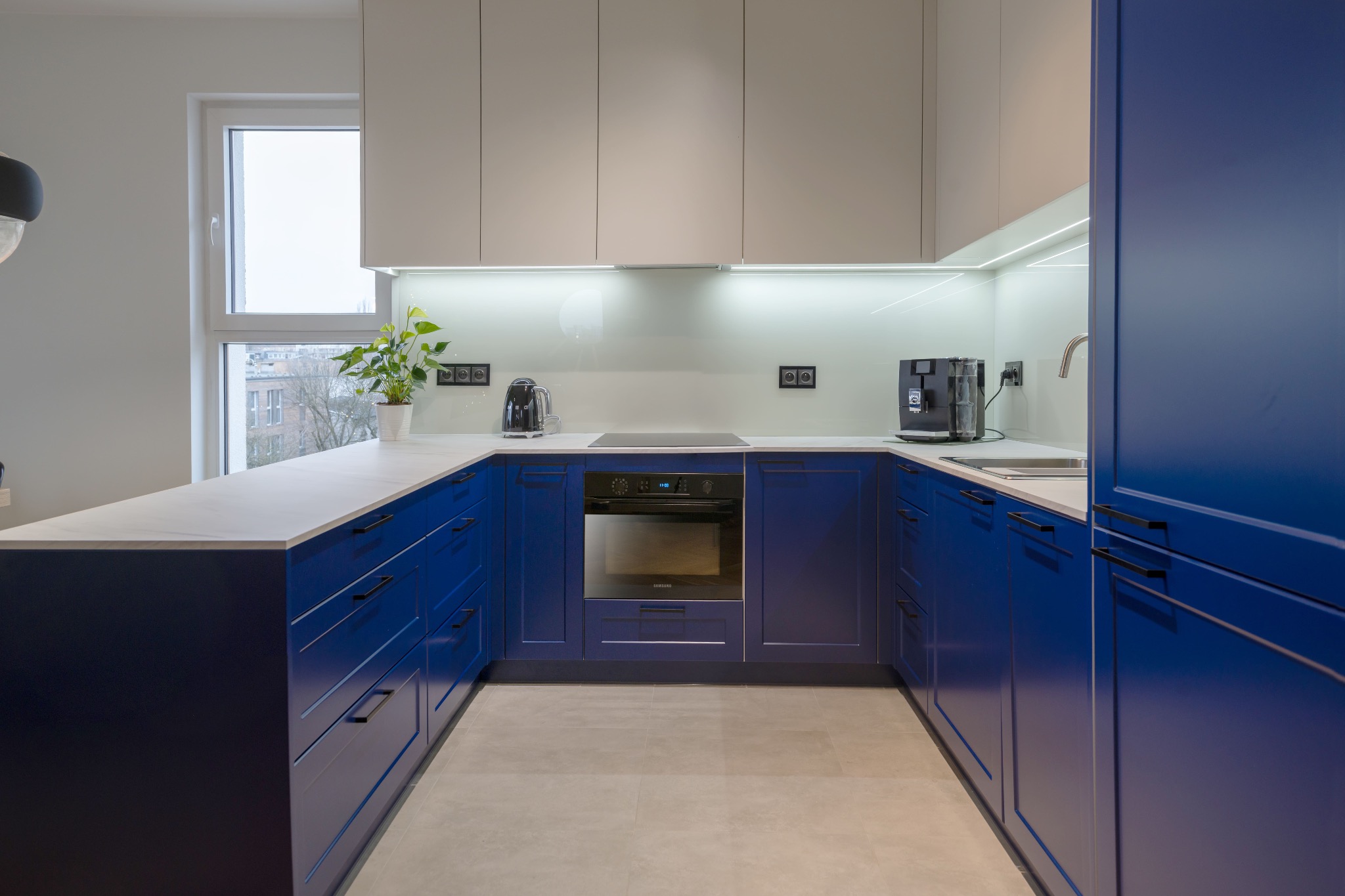 Nowoczesna kuchnia w kształcie litery U z niebieskimi szafkami i białym blatem. Nad blatem białe szafki wiszące i podświetlenie LED. Widok na okno z rośliną.