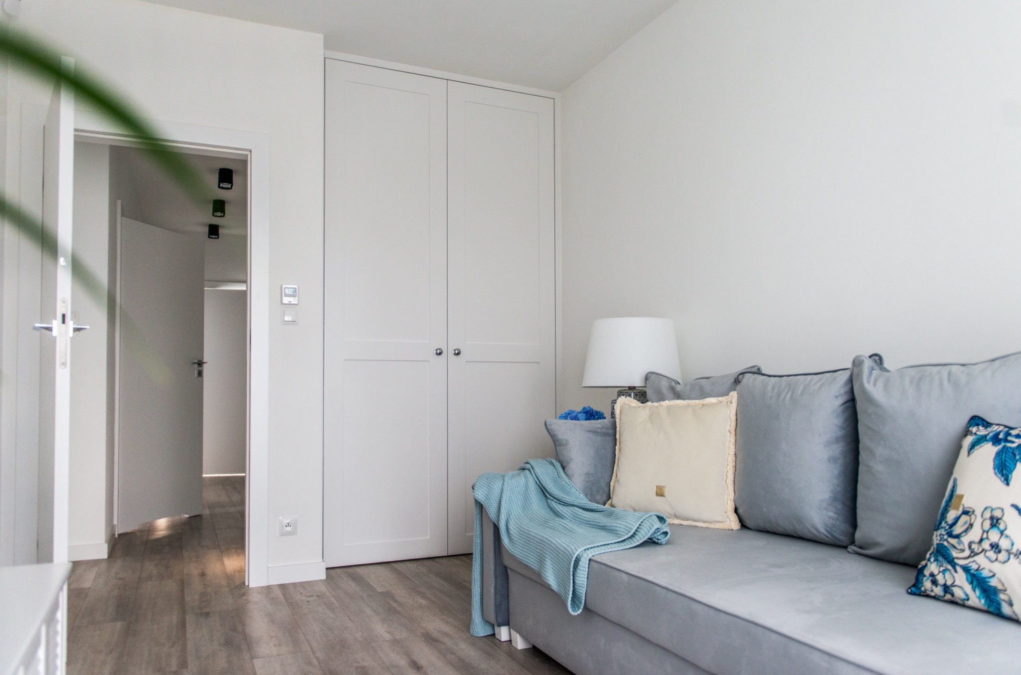Biała szafa wnękowa z dwoma drzwiami, widoczna z boku szara sofa z poduszkami i pledem. W tle otwarte drzwi do kolejnego pomieszczenia. Minimalistyczny wystrój.