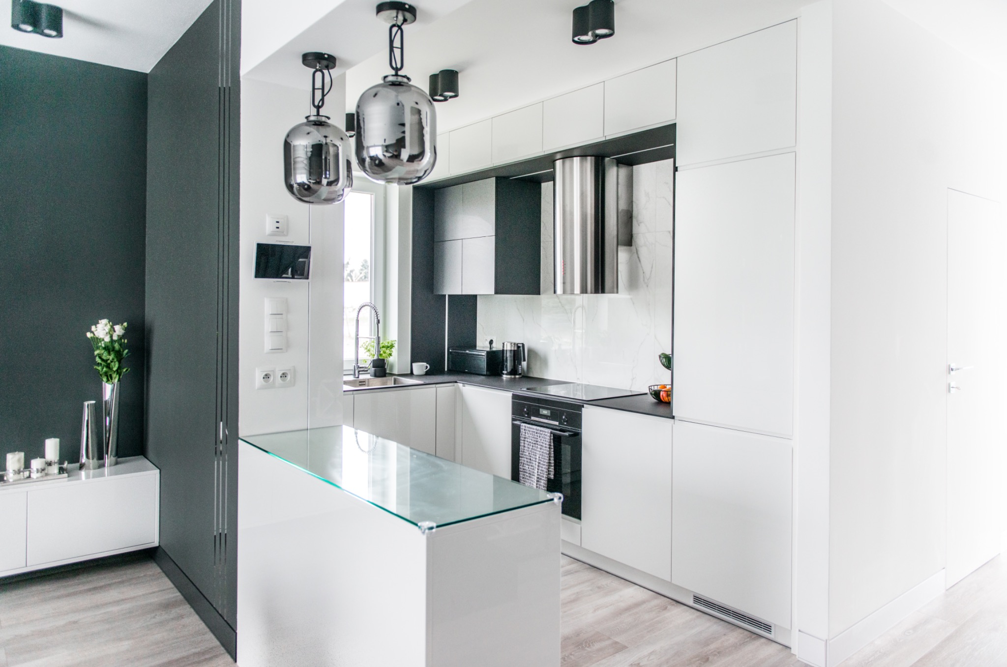 Nowoczesna, minimalistyczna kuchnia w bieli i szarości z wyspą z blatem ze szkła. Chromowane lampy wiszące i okap. Jasne drewniane panele podłogowe.