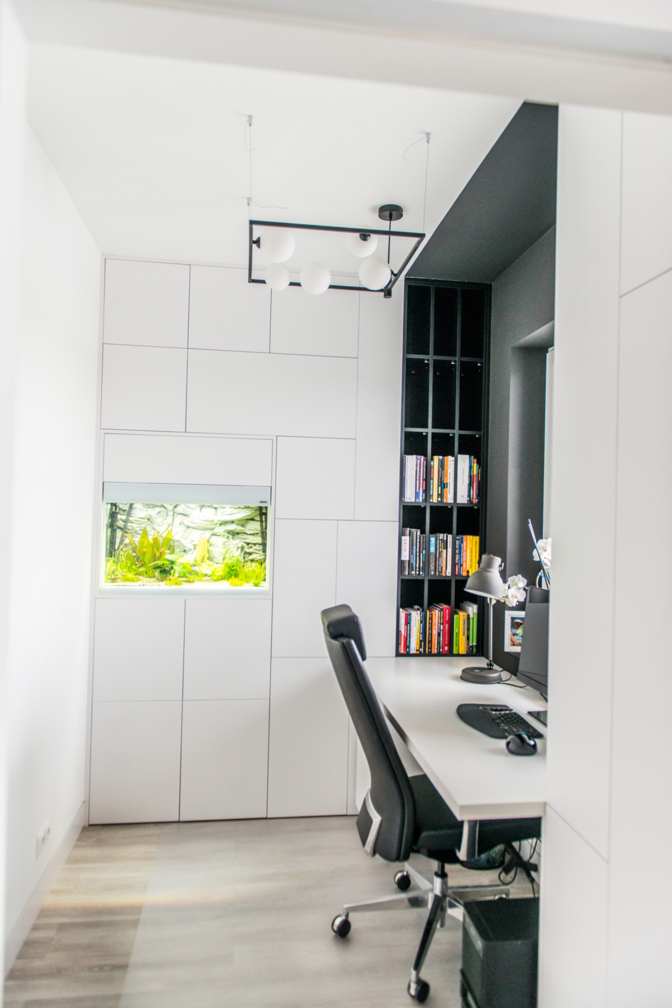 Nowoczesny pokój biurowy z białymi szafami w zabudowie, akwarium i czarnym regałem na książki. Minimalistyczne oświetlenie i biurko z krzesłem.