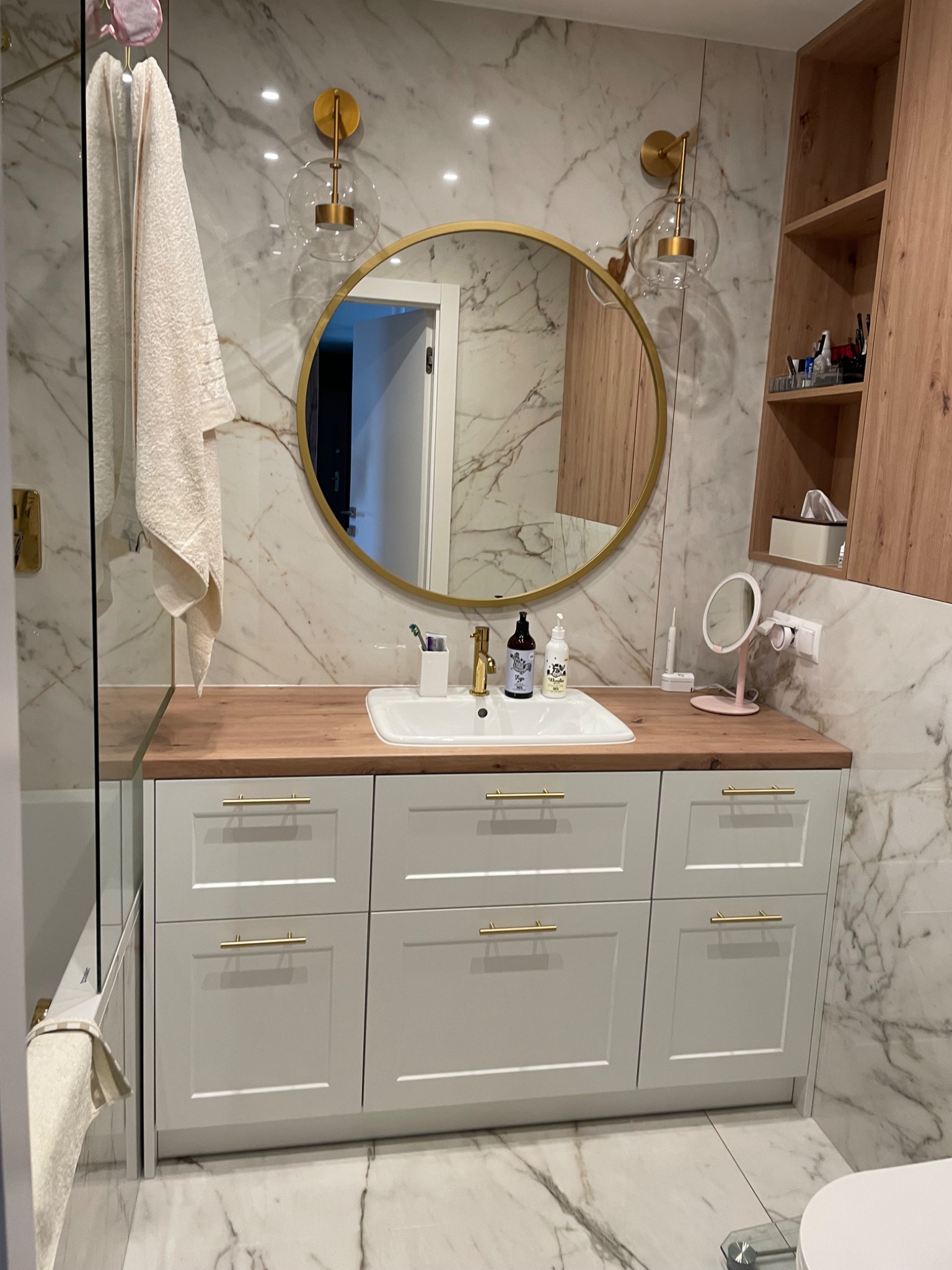 Jasna łazienka z marmurowymi płytkami, okrągłym lustrem w złotej ramie, drewnianym blatem i białą szafką z złotymi uchwytami. Nowoczesne oświetlenie.