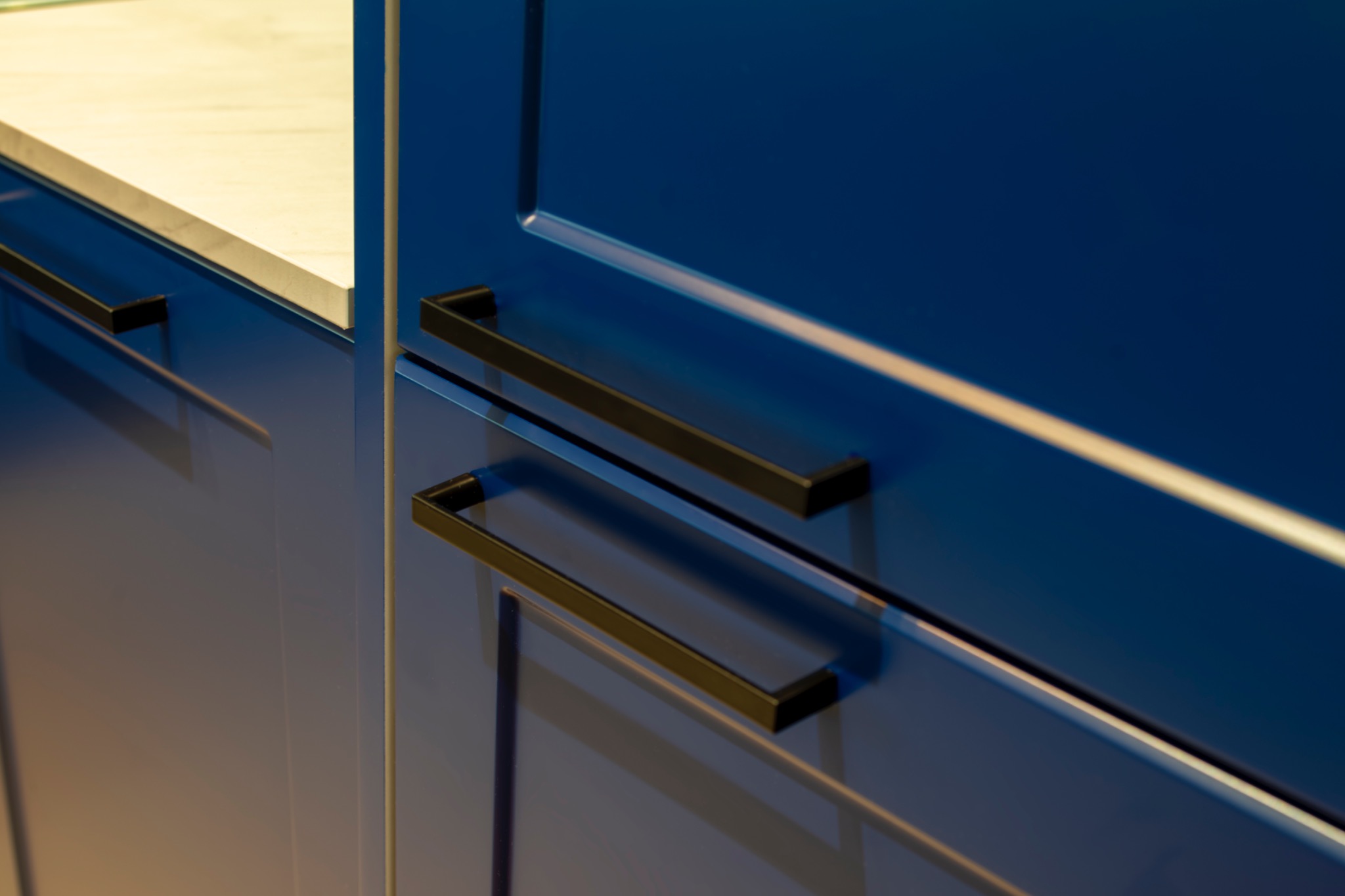 Niebieskie, lakierowane fronty szafek kuchennych z czarnymi, prostokątnymi uchwytami. Widoczny fragment blatu z jasnego kamienia. Nowoczesny design, gładkie powierzchnie.