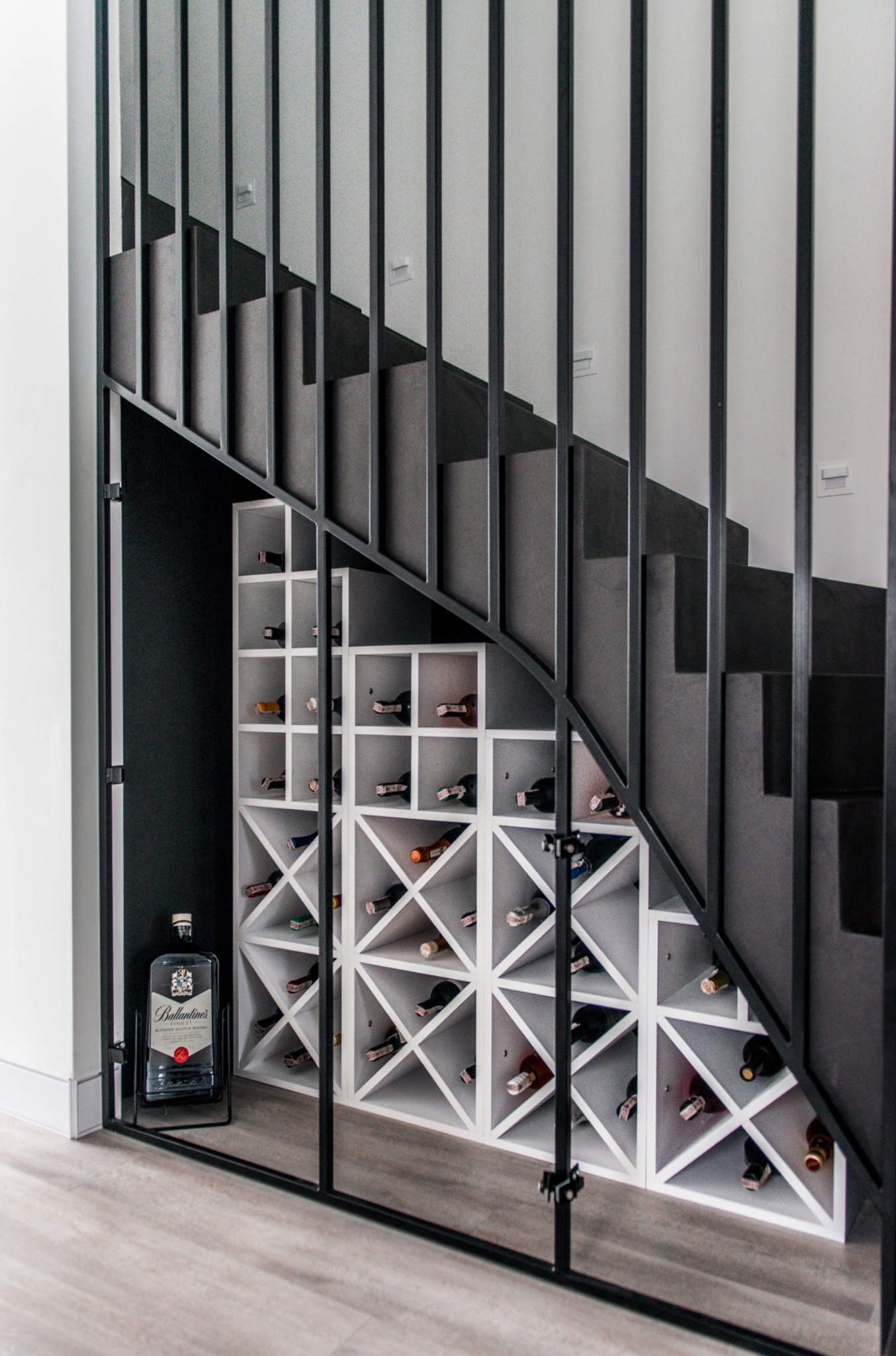 Biały regał na wino, wbudowany pod schodami z czarną balustradą. Na podłodze stoi butelka alkoholu. Nowoczesny design, funkcjonalne wykorzystanie przestrzeni.