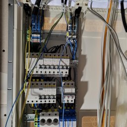 WOJCIECH ZIELEŃ - Rozdzielnica elektryczna z bezpiecznikami i przewodami, widok z bliska. Elementy instalacji w szarej obudowie, kable w różnych kolorach.