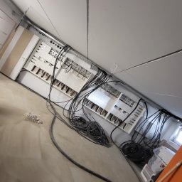 WOJCIECH ZIELEŃ - Rozdzielnia elektryczna w trakcie montażu: widoczne kable, listwy zaciskowe i elementy instalacji w surowym wnętrzu. Ujęcie z nietypowej perspektywy.