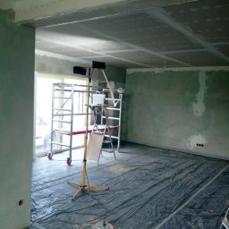 Pasjograf - Montaż płyt gipsowo-kartonowych na suficie w trakcie prac wykończeniowych. Widoczne rusztowanie, lampa oświetleniowa i folia ochronna na podłodze.