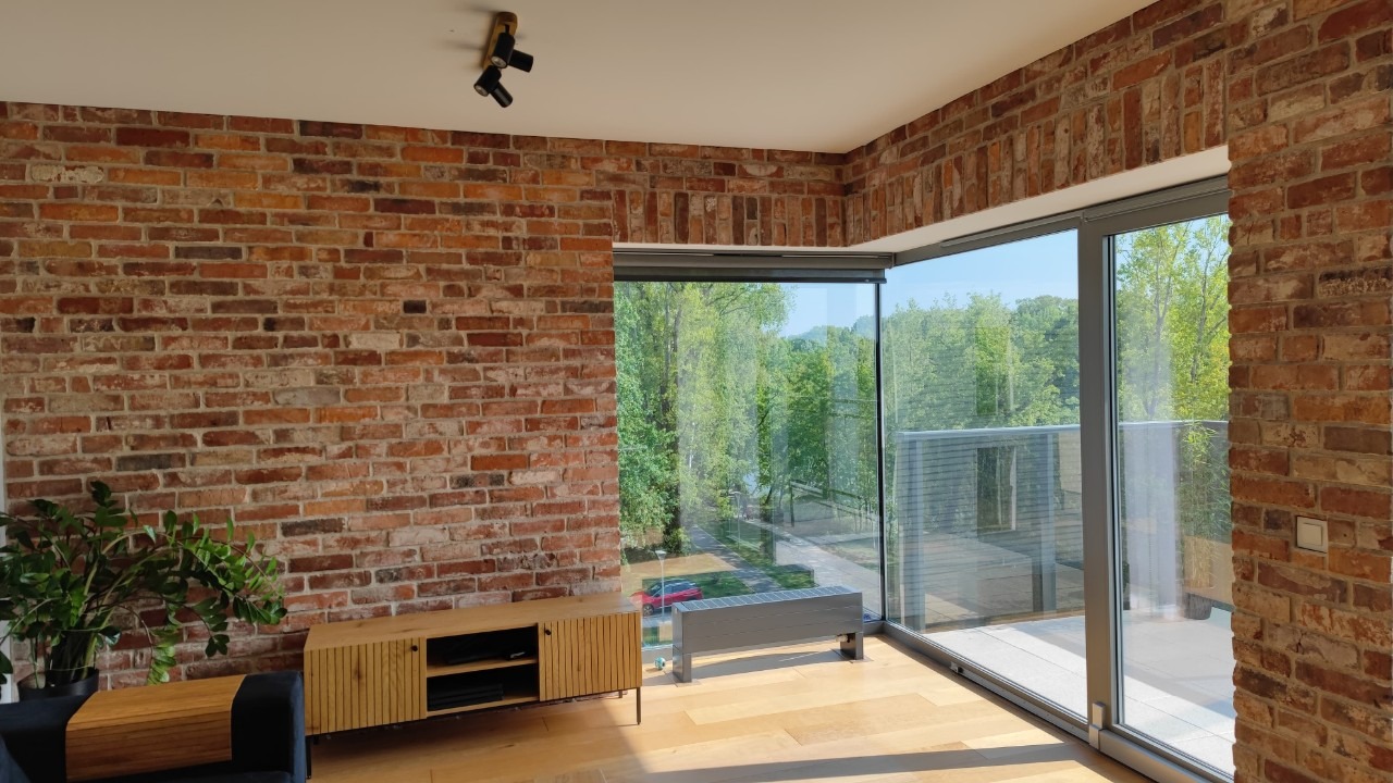 Salon z ceglaną ścianą, dużym oknem z widokiem na zieleń i drewnianą podłogą. Widoczny fragment sofy i szafka RTV. Oświetlenie punktowe na suficie.