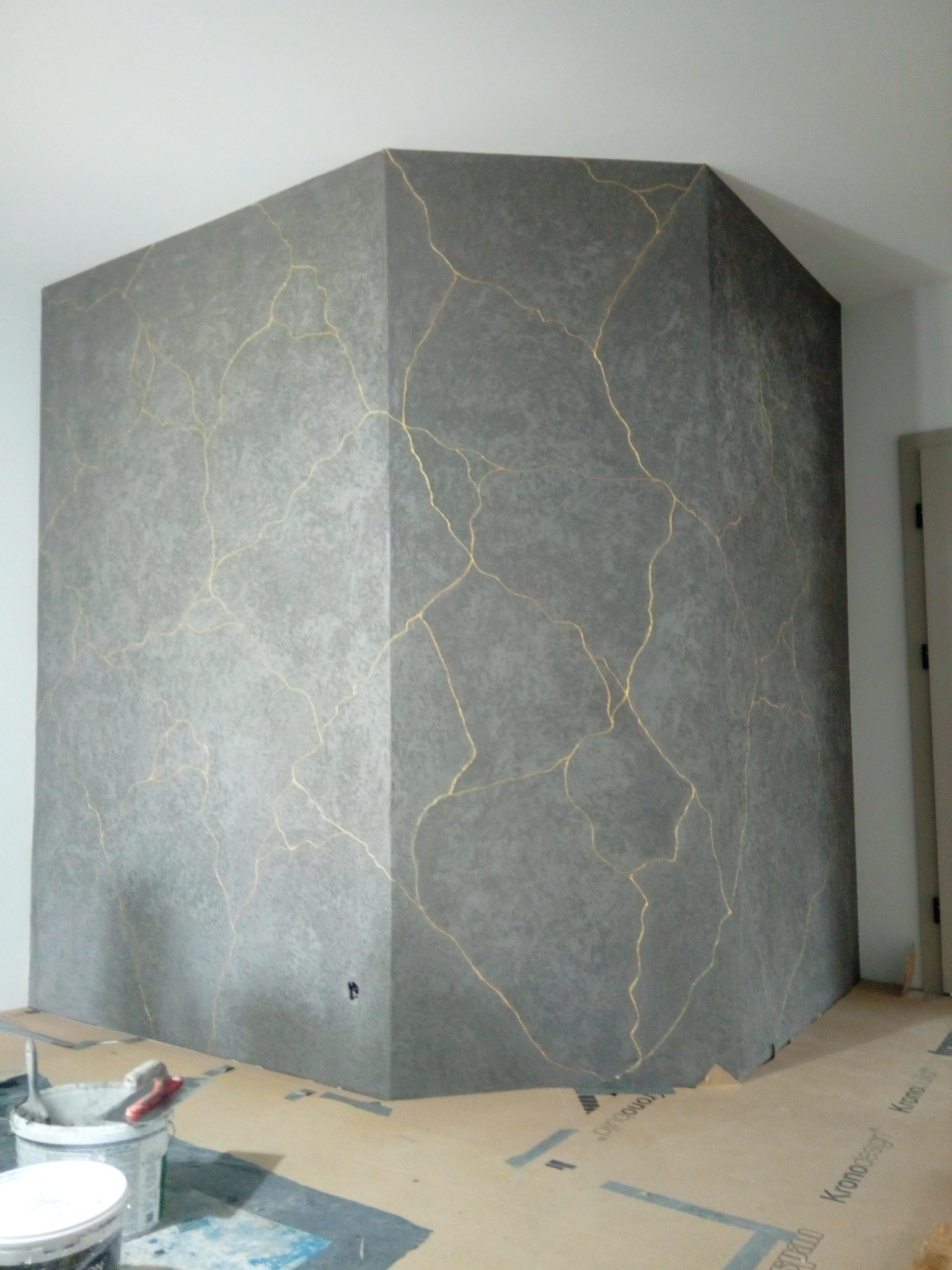 Dekoracyjna ściana w odcieniach szarości z efektem złotych pęknięć, wykonana techniką Kintsugi. Widoczne narzędzia malarskie i zabezpieczona podłoga.