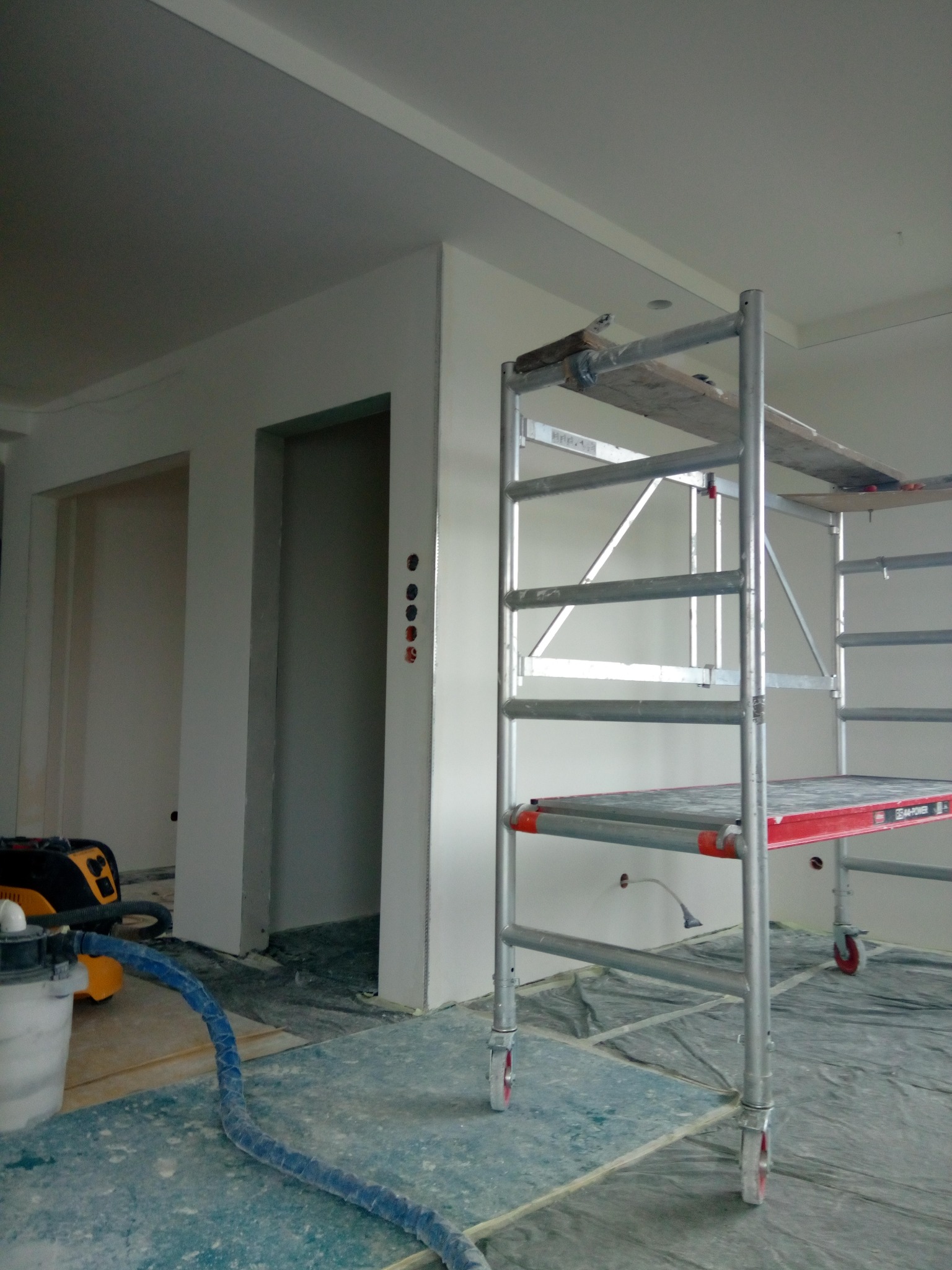 Białe ściany w trakcie prac wykończeniowych, widoczne rusztowanie, szlifierka i odkurzacz przemysłowy. Przygotowanie do malowania.