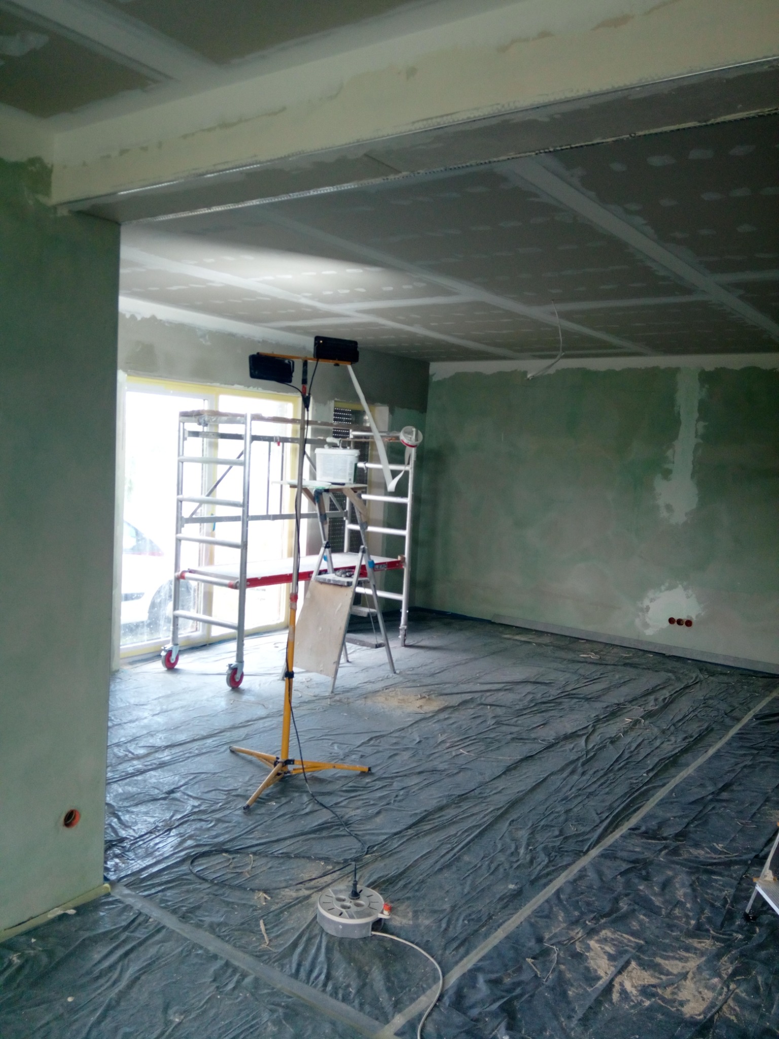 Montaż płyt gipsowo-kartonowych na suficie w trakcie prac wykończeniowych. Widoczne rusztowanie, lampa oświetleniowa i folia ochronna na podłodze.