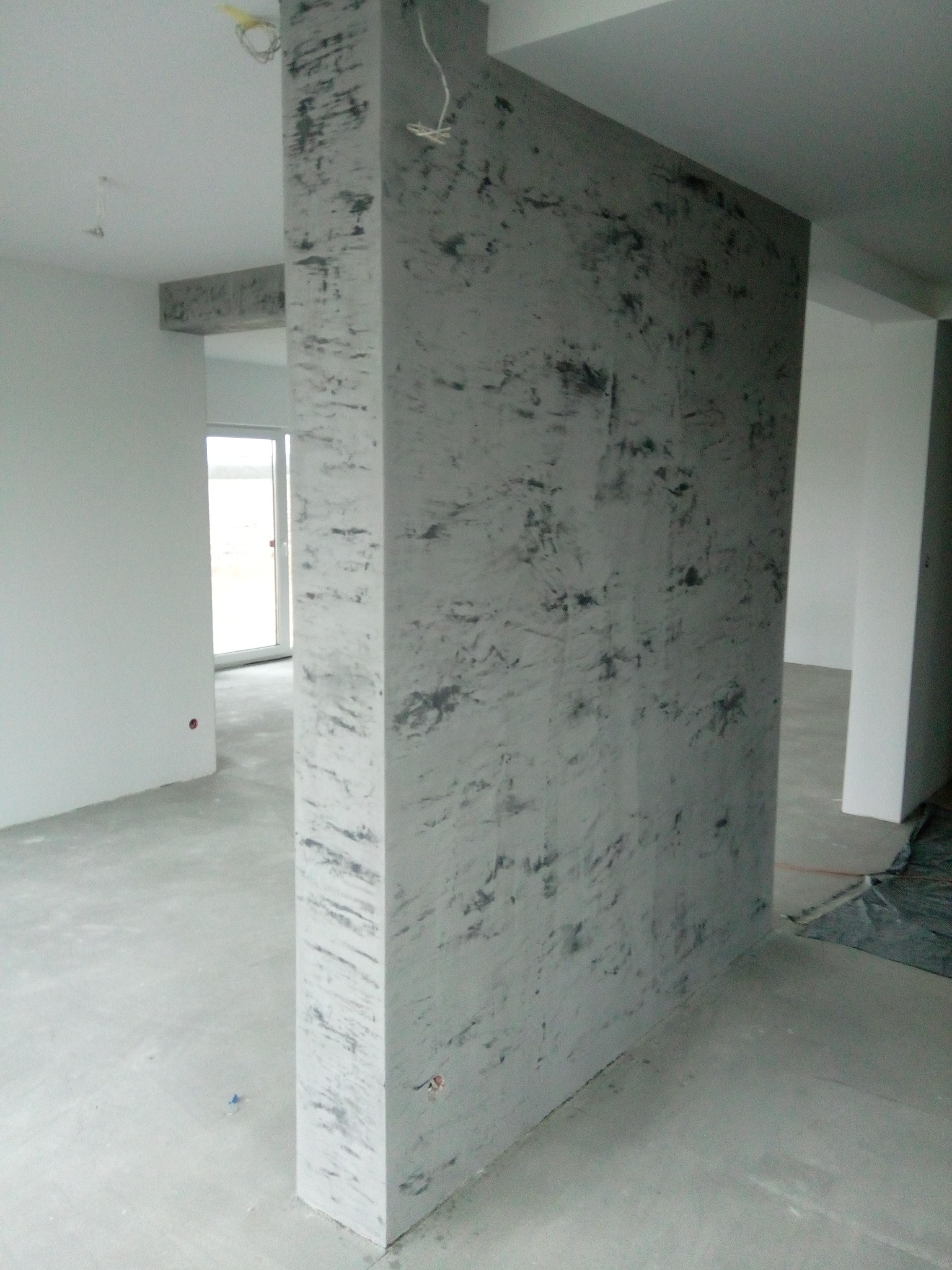 Ściana działowa z efektem dekoracyjnym w szarościach, widoczna w surowym wnętrzu. Element wykończenia wnętrza, z widocznymi przewodami i oknem w tle.