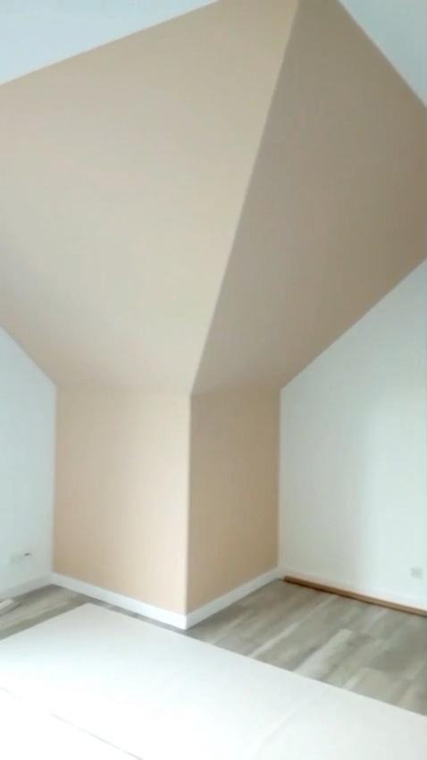 Pomieszczenie po malowaniu z nietypowym załamaniem sufitu, ściany w odcieniach beżu i bieli, szara podłoga. Widok na róg pokoju.