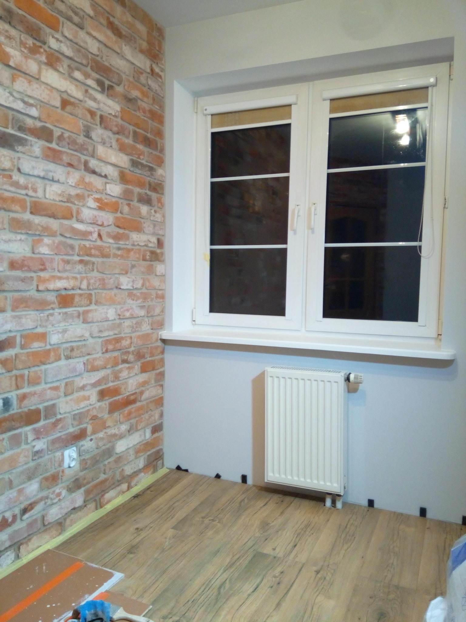 Pokój po remoncie: ściana z cegły dekoracyjnej, drewniana podłoga, białe okno z roletami i grzejnik. Widoczne narzędzia i materiały na podłodze.