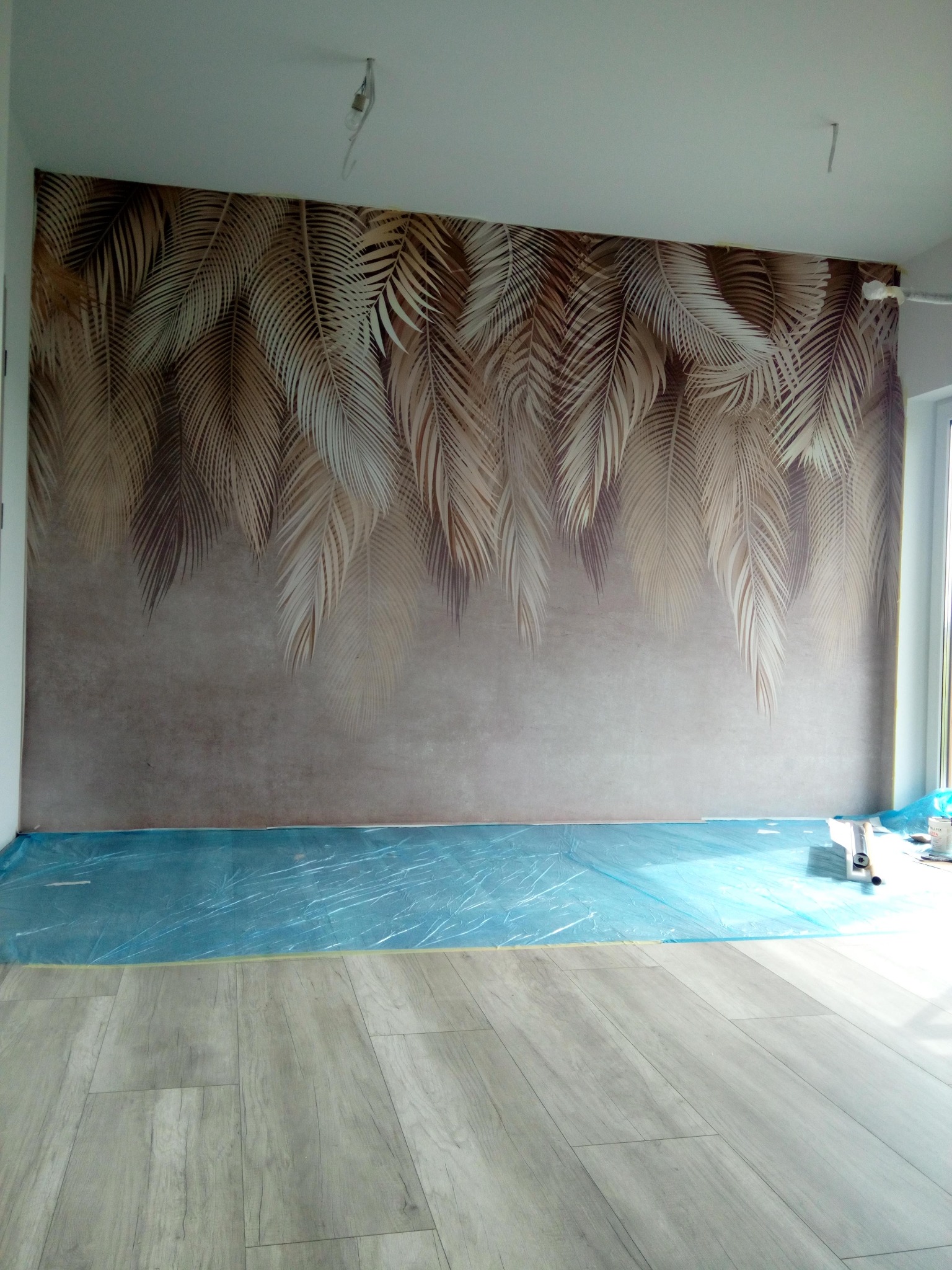 Pokój po tapetowaniu z motywem liści palmowych, podłoga zabezpieczona folią. Widoczne narzędzia i resztki kleju. Jasne panele podłogowe.
