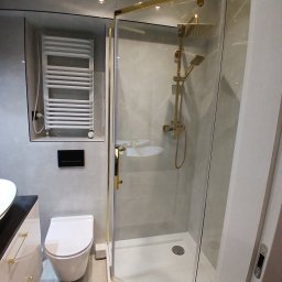 Volodymyr Mazepa - Nowoczesna łazienka z kabiną prysznicową w złotych ramach, wiszącą toaletą i grzejnikiem drabinkowym. Minimalistyczny design, szare płytki i białe elementy wyposażenia.