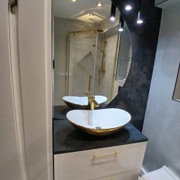 Volodymyr Mazepa - Nowoczesna łazienka z owalną umywalką w kolorze białym ze złotymi akcentami, czarnym blatem i białymi szafkami. W tle widoczne lustro i kabina prysznicowa.