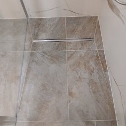 Volodymyr Mazepa - Ukończona podłoga w kabinie prysznicowej z odpływem liniowym. Płytki imitujące marmur w odcieniach beżu i szarości. Widoczny fragment kabiny i baterii prysznicowej.