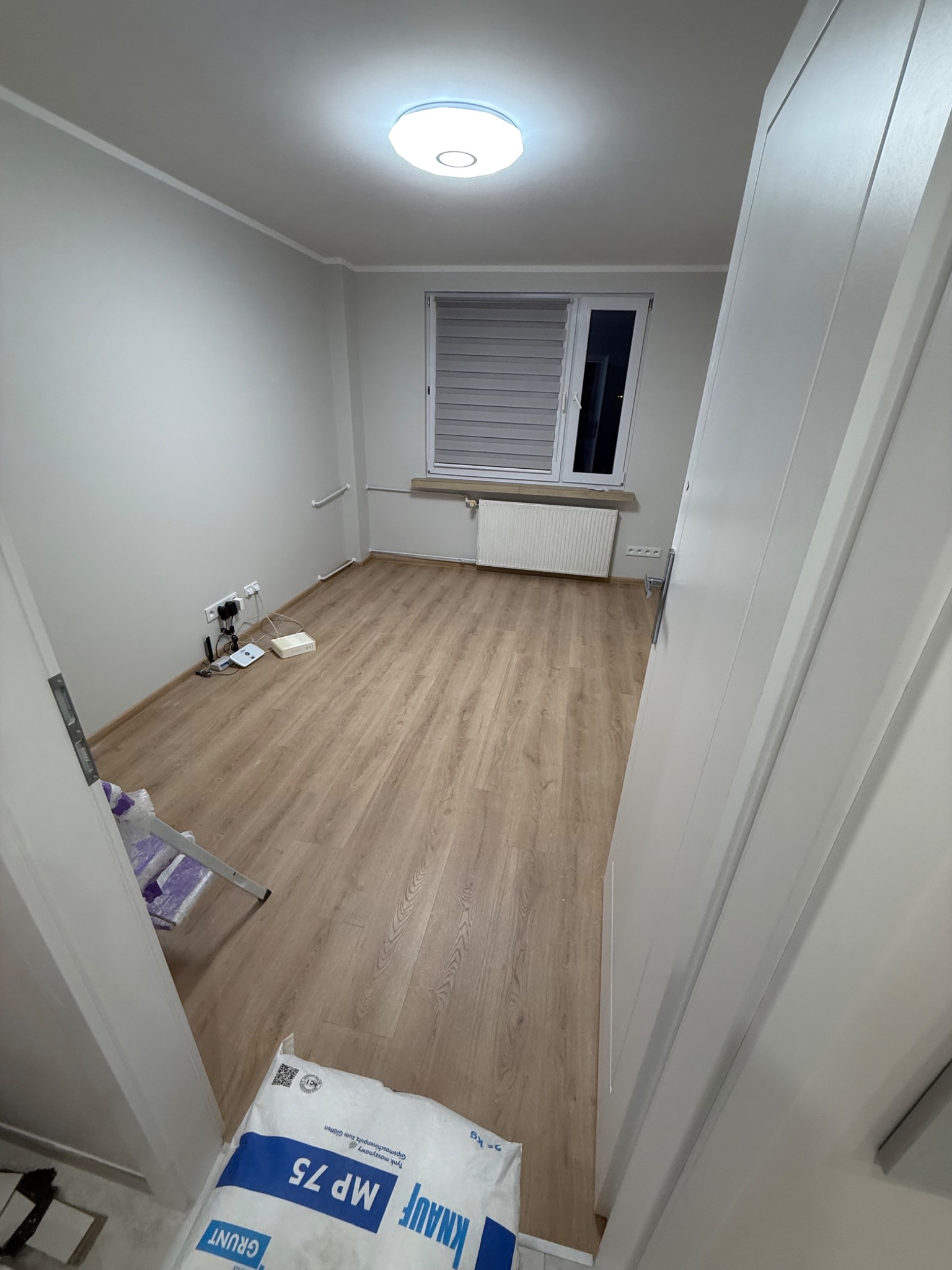 Jasne wnętrze pokoju po remoncie z drewnopodobną podłogą, grzejnikiem pod oknem i workiem gładzi Knauf MP 75. Widok zza otwartych drzwi.