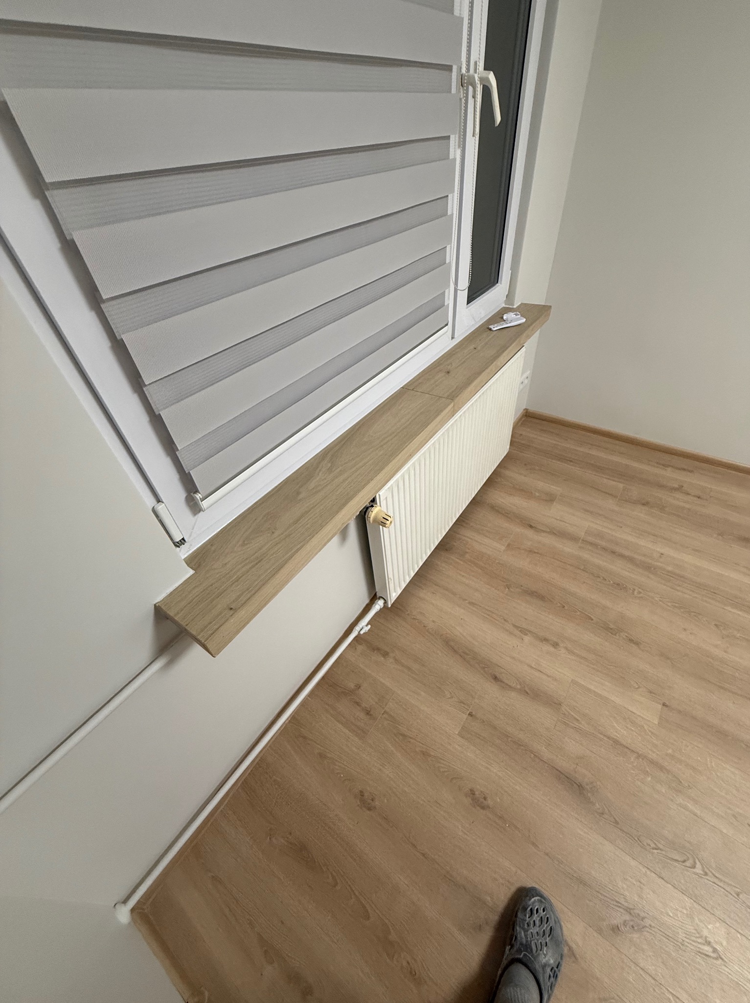 Biały grzejnik z drewnianym blatem pod oknem z roletami dzień-noc, na tle jasnych ścian i podłogi z paneli. Widoczny fragment stopy w obuwiu.