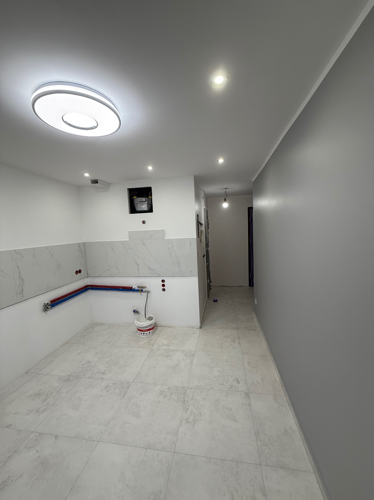 Wnętrze kuchni w trakcie prac wykończeniowych: ułożone płytki, częściowo położona glazura, widoczne rury instalacji wodnej. Ściana pomalowana na szaro.