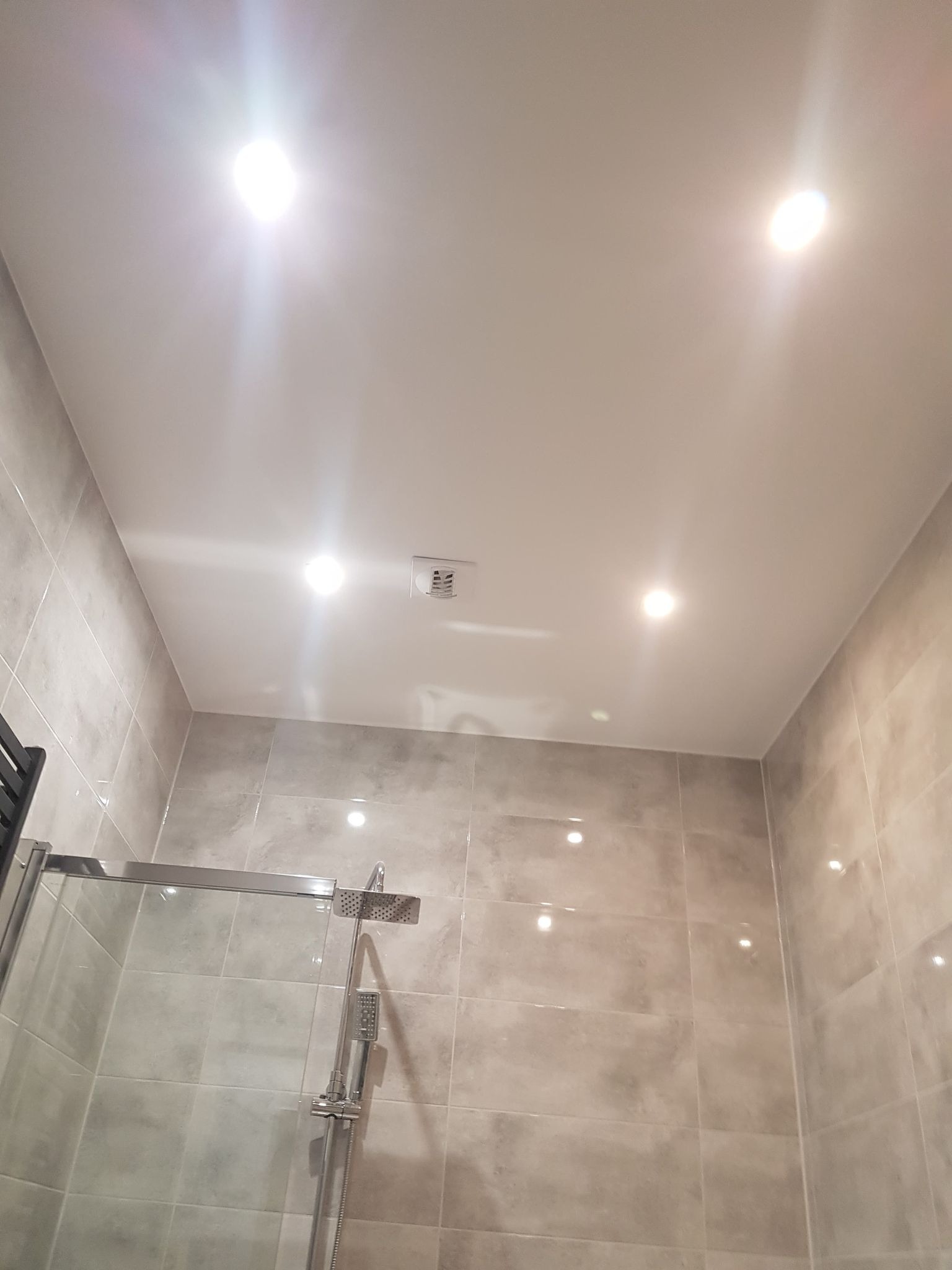 Wykończona łazienka z beżowymi płytkami, kabiną prysznicową ze szklanymi drzwiami i punktowym oświetleniem sufitowym. Widok z dołu, podkreślający detale wykończenia.