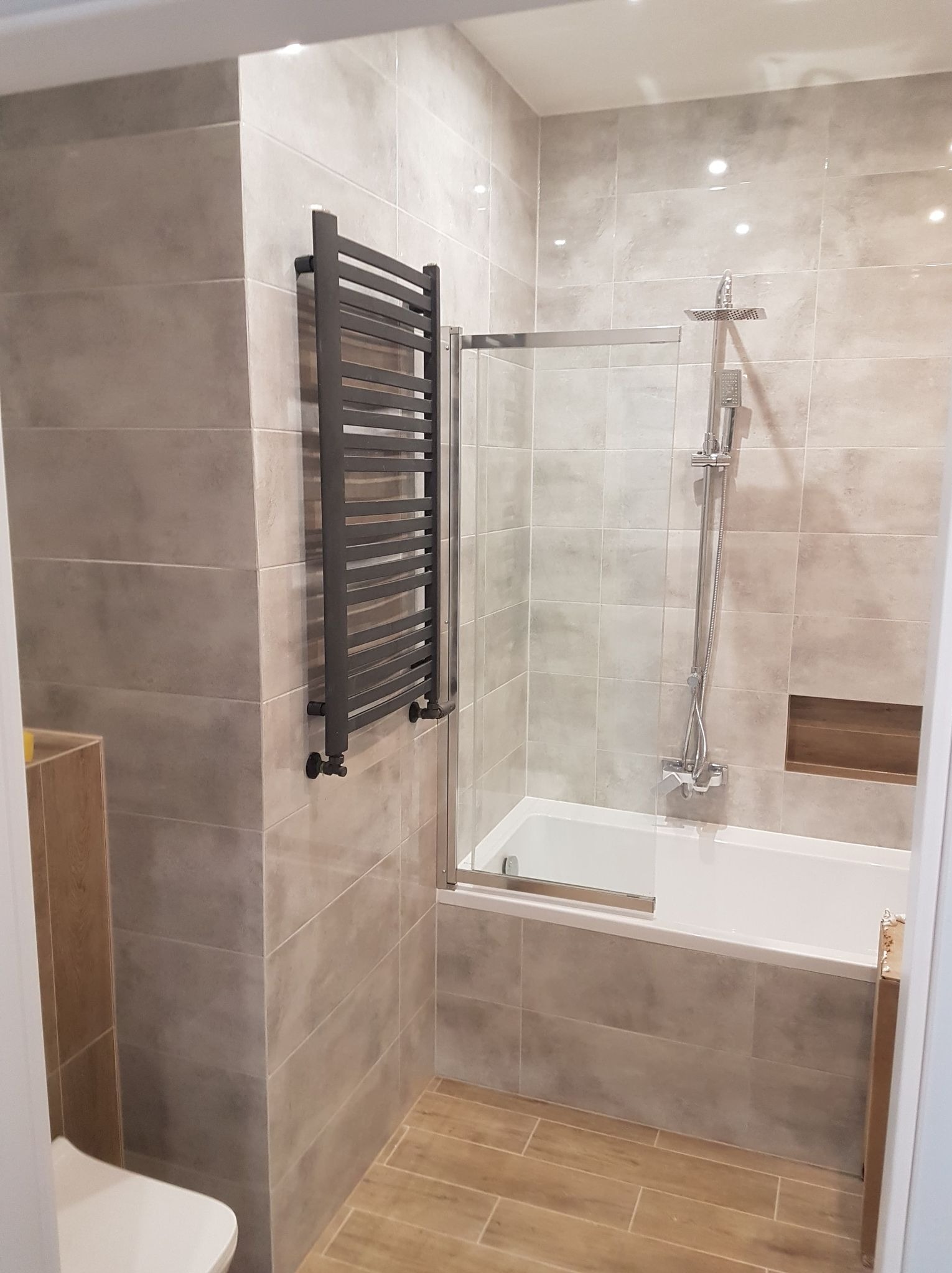 Wykończona łazienka z beżowymi płytkami, kabiną prysznicową nad wanną i grzejnikiem drabinkowym. Widoczna drewnopodobna podłoga i wnęka w ścianie.