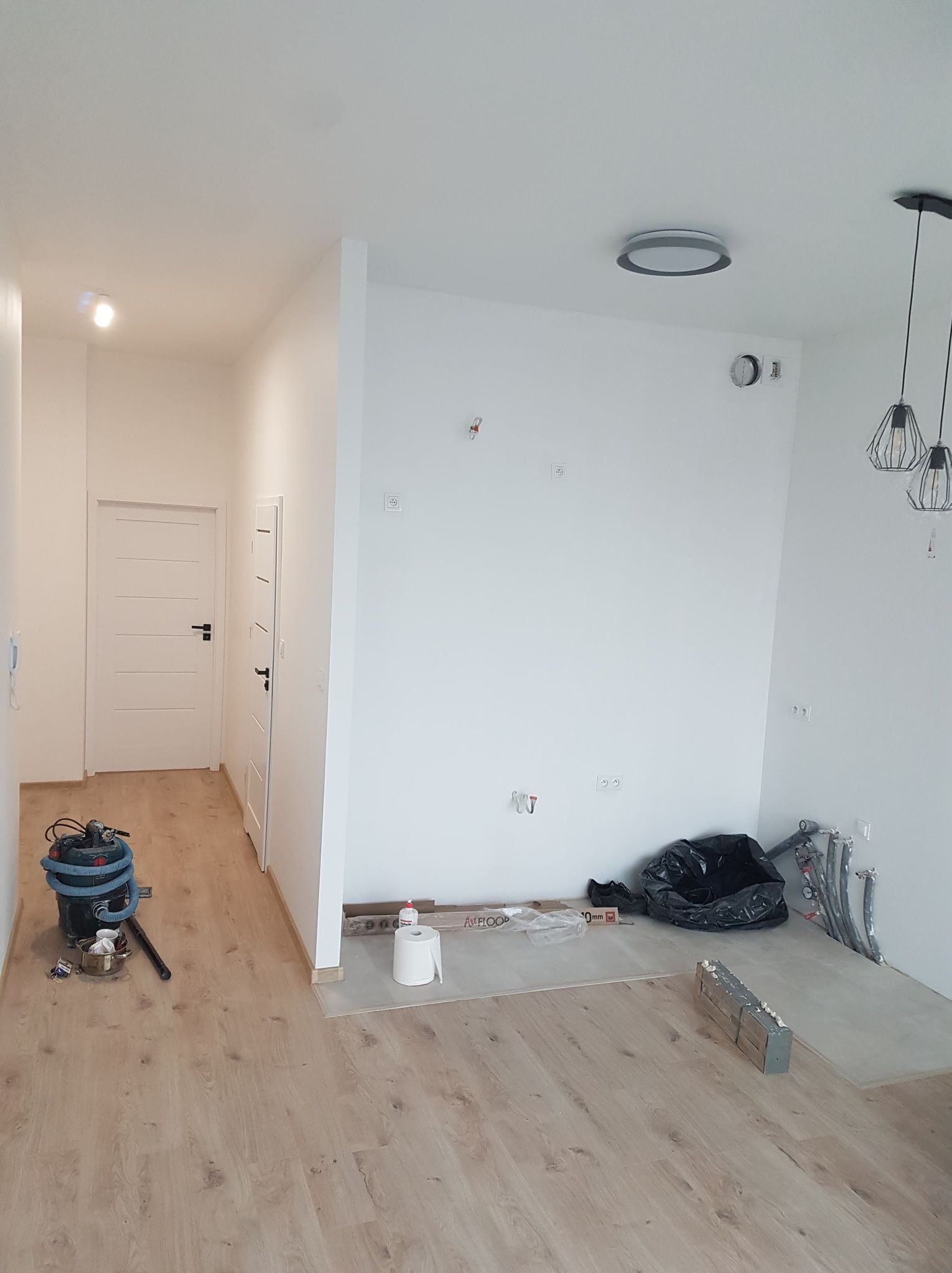 Jasne wnętrze w trakcie prac wykończeniowych: białe ściany, drewnopodobna podłoga, widoczne instalacje, narzędzia i materiały budowlane. Minimalistyczne lampy sufitowe.