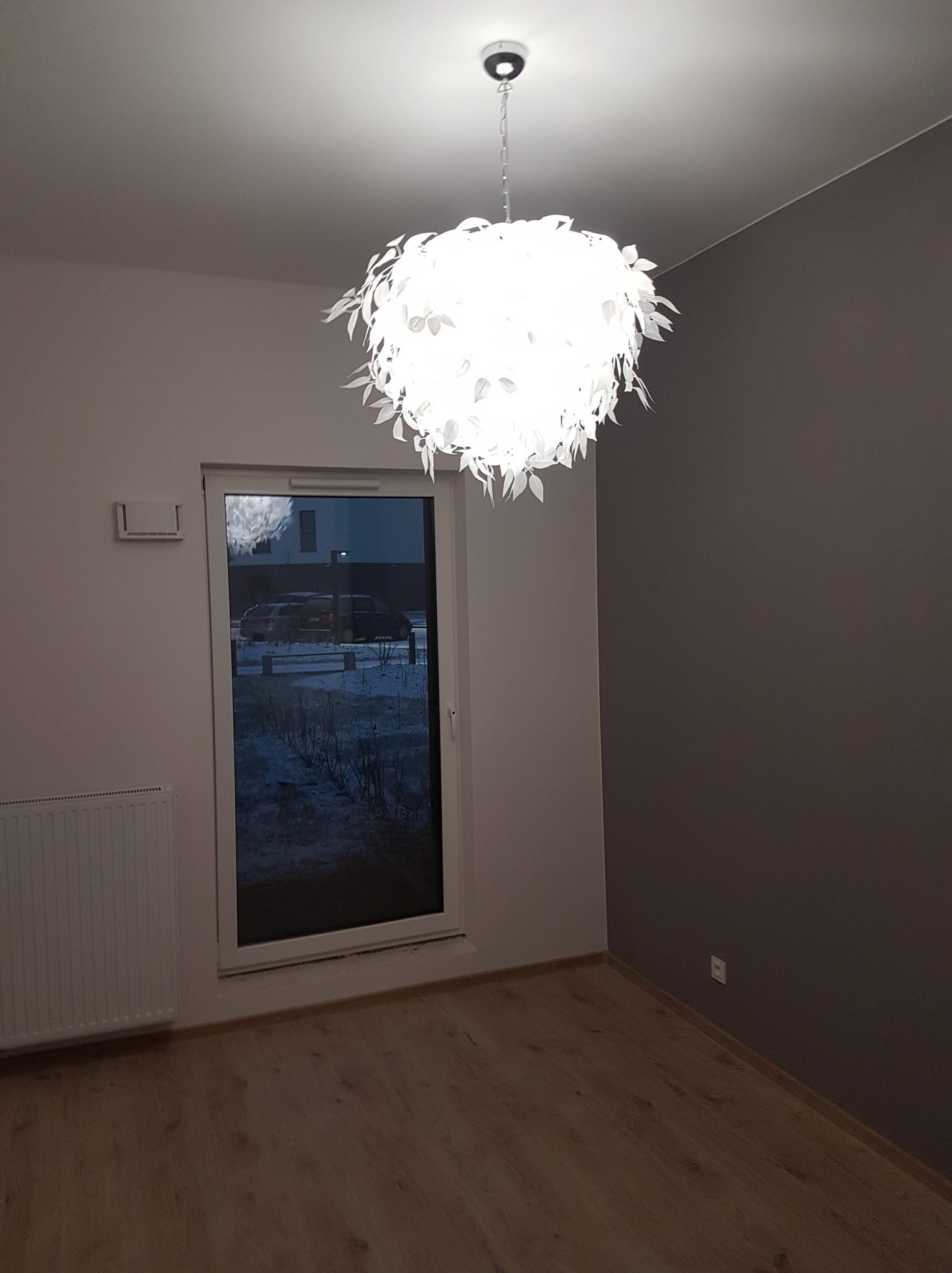 Pomalowane na szaro wnętrze pokoju z drewnianą podłogą, białym grzejnikiem i oknem. W centrum ozdobny, biały żyrandol w kształcie kuli z elementami przypominającymi liście.