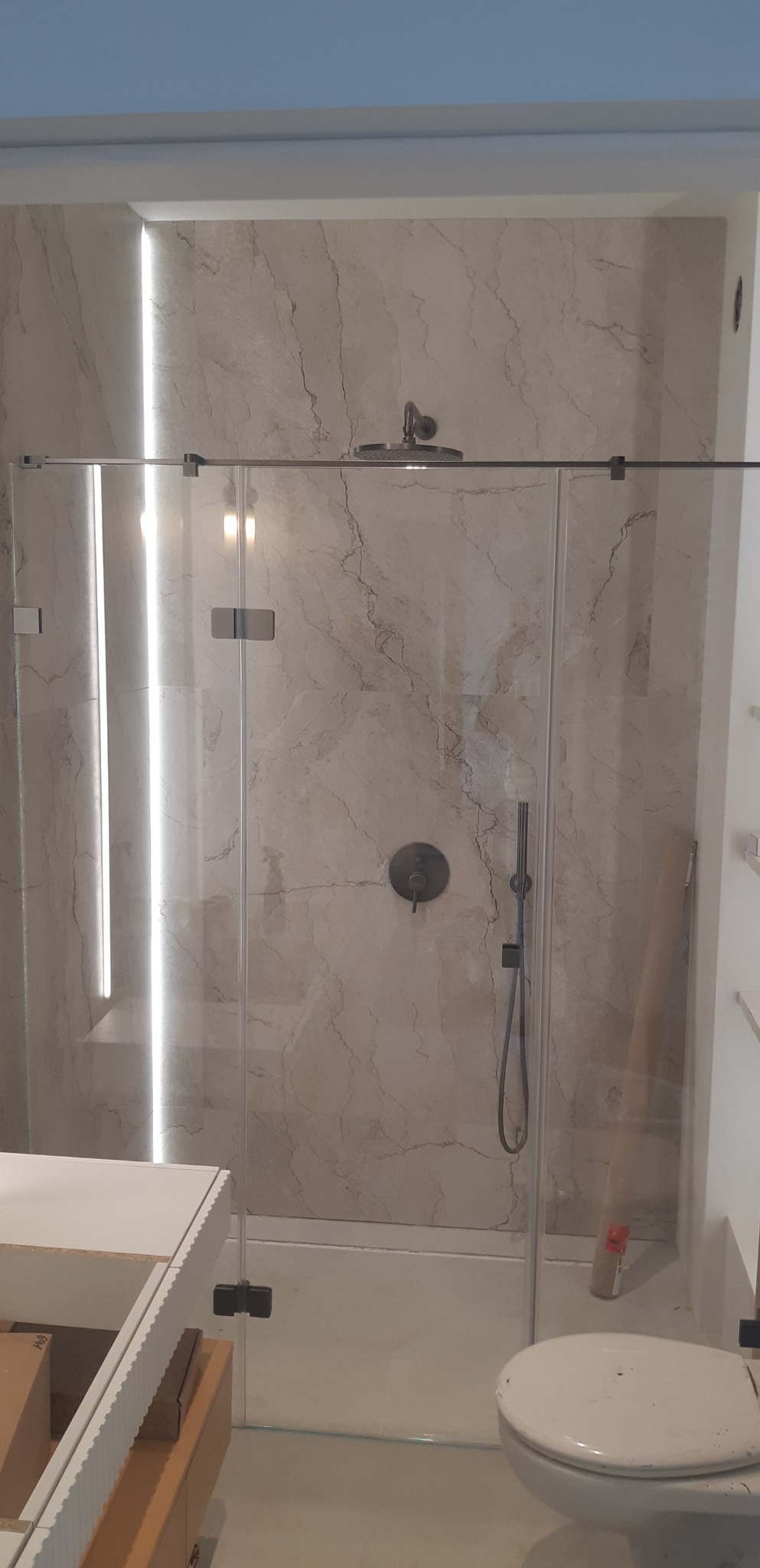 Wykończona łazienka z kabiną prysznicową typu walk-in, marmurowe płytki na ścianach, oświetlenie LED. Widoczna toaleta i fragment szafki pod umywalkę.