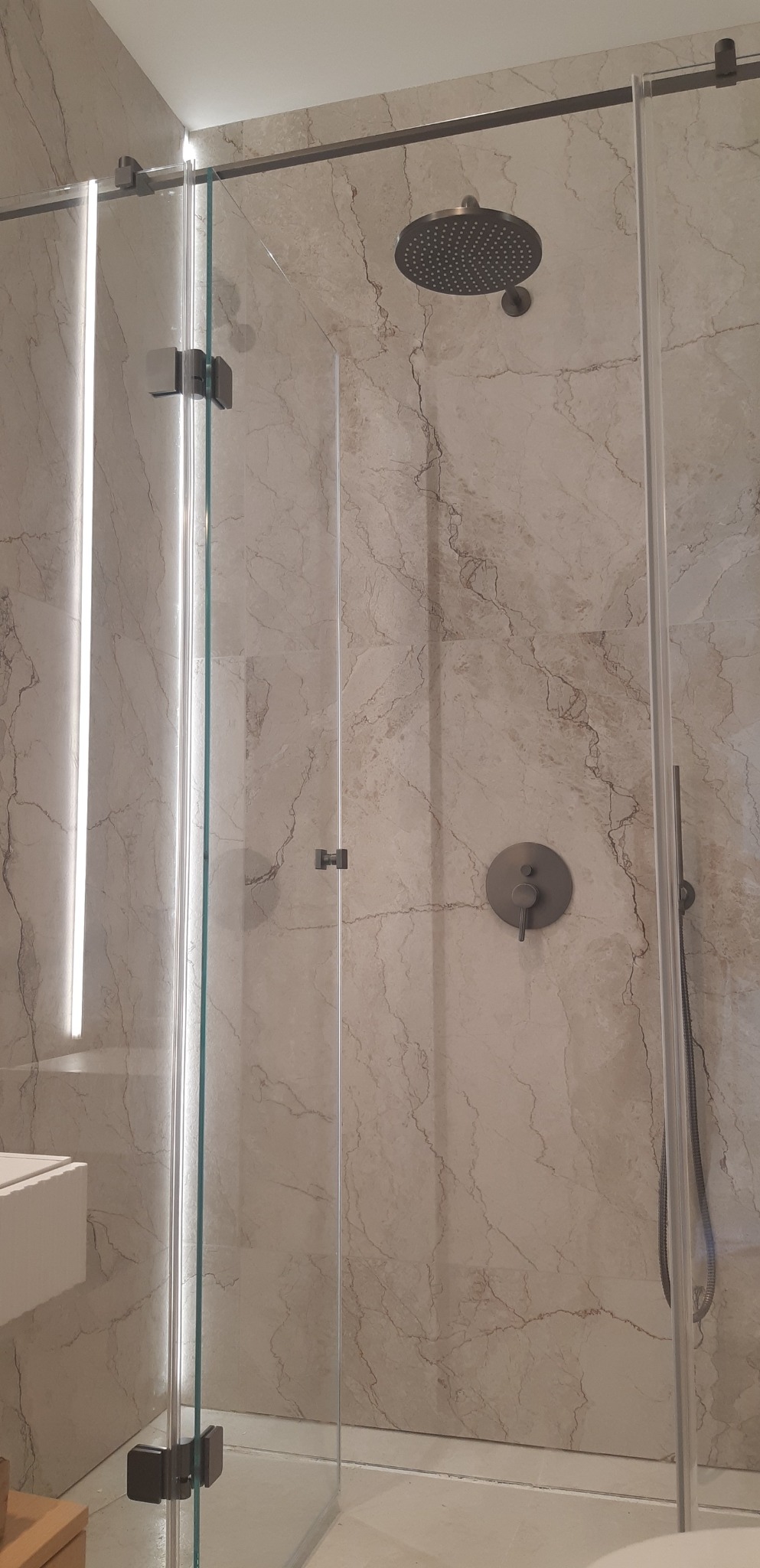 Nowoczesna kabina prysznicowa z deszczownicą i szklanymi drzwiami przesuwnymi, wykończona marmurową płytką w beżowych odcieniach. Minimalistyczny design.