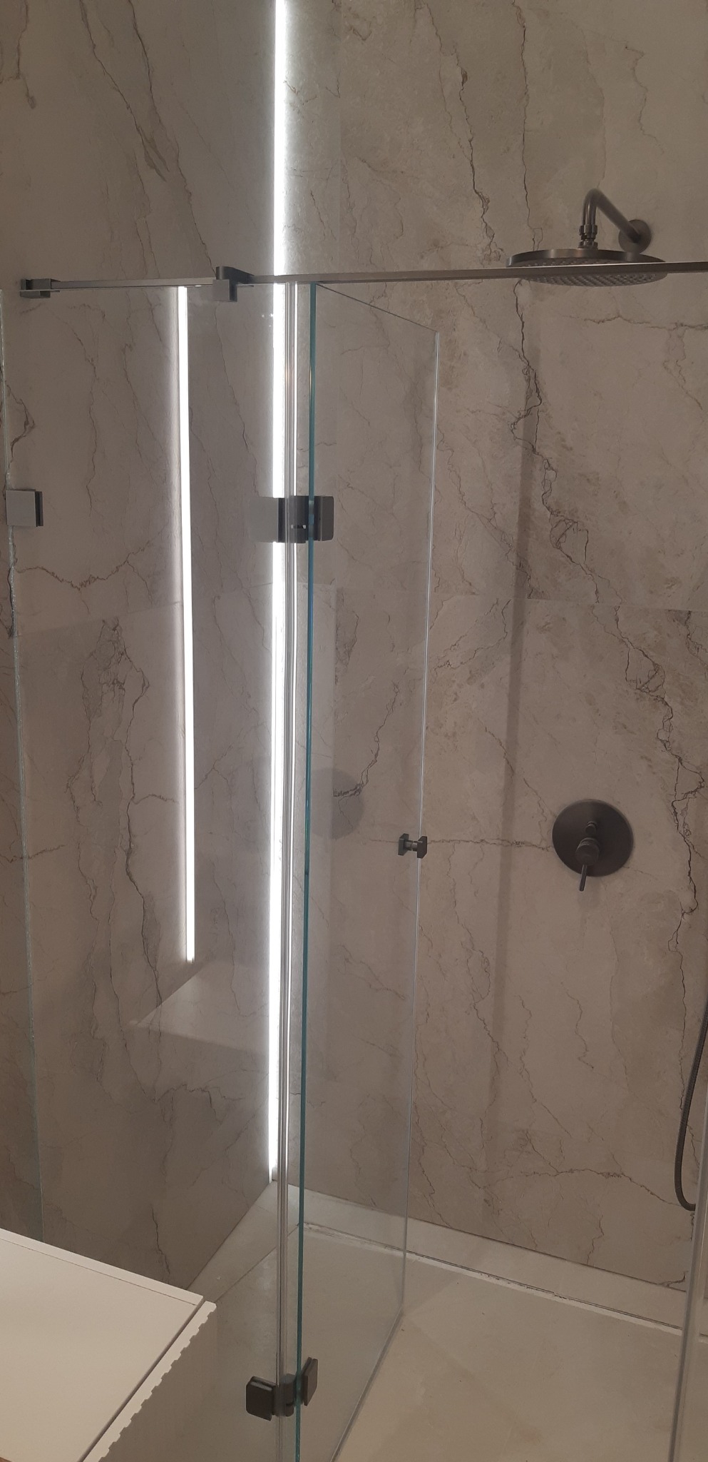 Nowoczesna kabina prysznicowa z szklanymi drzwiami i deszczownicą, wykończona płytkami imitującymi marmur z subtelnym oświetleniem LED.