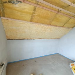 Rolling Tubes Bartłomiej Chmielewski - Ocieplenie poddasza wełną mineralną, widoczne drewniane belki konstrukcyjne i folia paroizolacyjna. Przy podłodze leżą rolki wełny i worki. Ściany pomalowane na biało.
