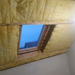 Rolling Tubes Bartłomiej Chmielewski - Izolacja poddasza wełną mineralną z oknem dachowym, zabezpieczona sznurkami. Widoczny fragment ściany i grzejnik. Prace wykończeniowe w trakcie realizacji.