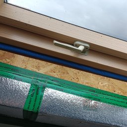 Rolling Tubes Bartłomiej Chmielewski - Montaż okna dachowego: widok na drewnianą ramę, uszczelnienie folią aluminiową Tesa Seal Flex Plus 60077 i niebieski przewód. Widoczna klamka okienna Fakro.