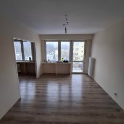 Rolling Tubes Bartłomiej Chmielewski - Jasne, przestronne wnętrze po remoncie z panelami podłogowymi, świeżo pomalowanymi ścianami i dużymi oknami z widokiem na zewnątrz. Wnętrze do wykończenia.