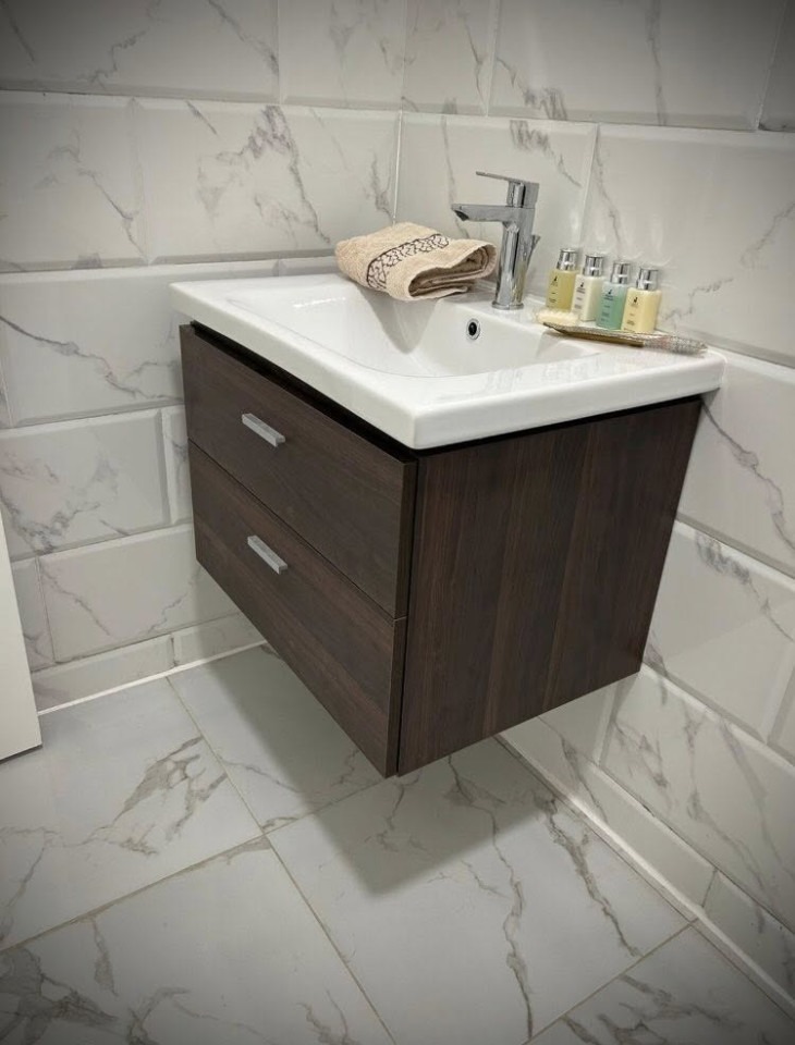 Nowoczesna łazienka: biała umywalka z ciemnobrązową szafką, marmurowe płytki na ścianach i podłodze. Na umywalce ręcznik i zestaw kosmetyków.