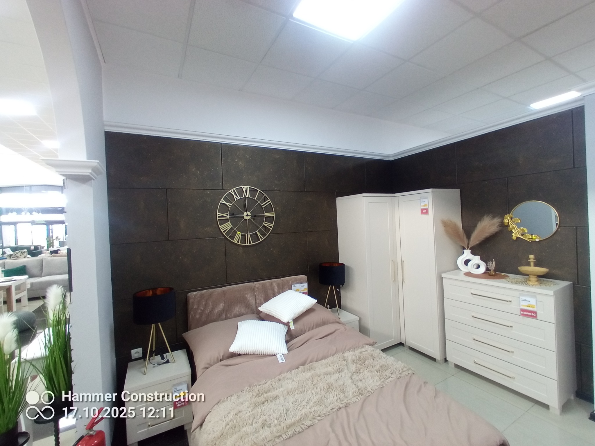 Nowoczesna sypialnia z łóżkiem, szafkami nocnymi, komodą i szafą w jasnych barwach. Na ścianie dekoracyjny zegar i lustro. Całość w minimalistycznym stylu.