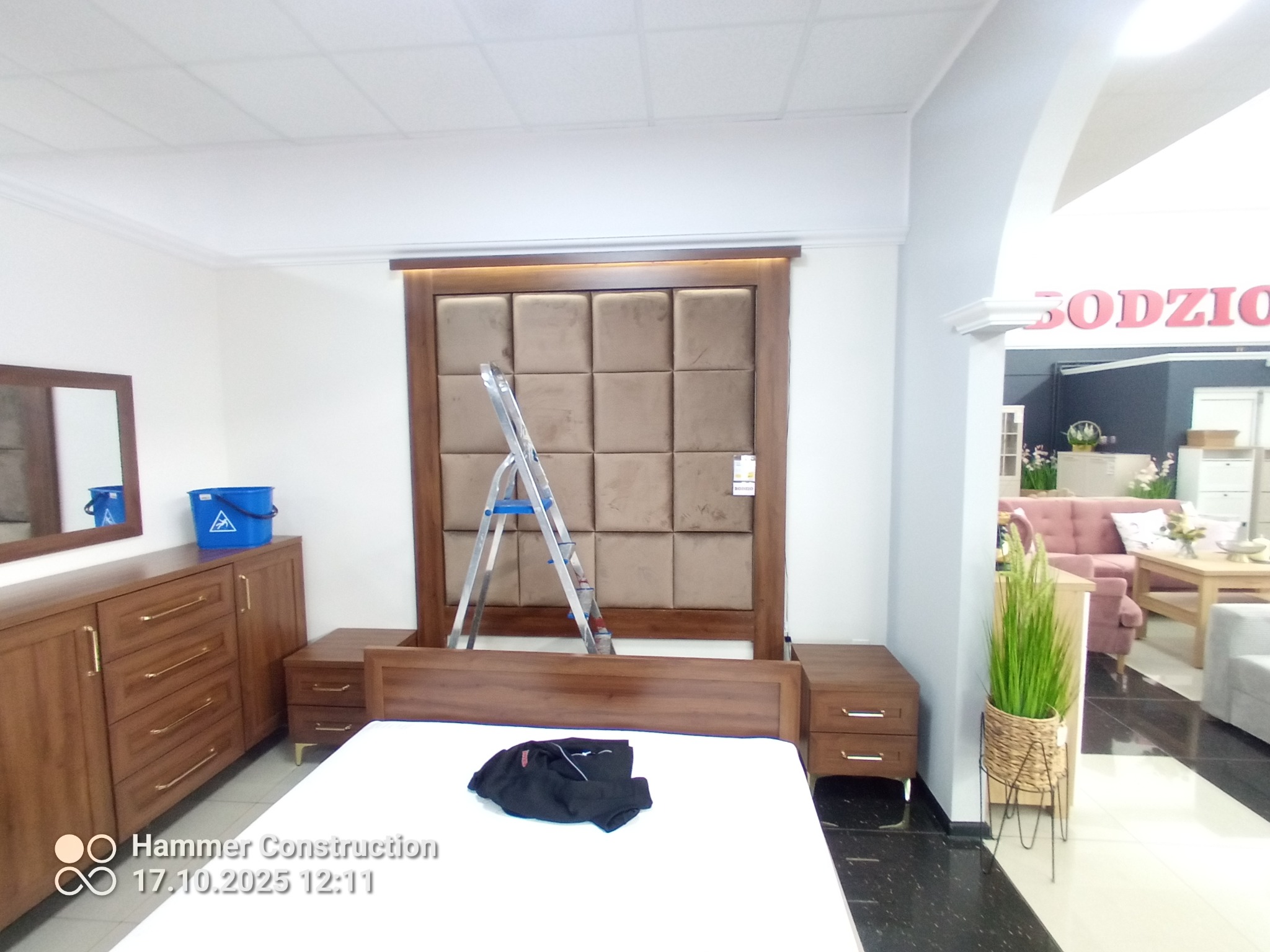 Tapicerowany zagłówek łóżka w brązach, z drabinką opartą o niego. Widoczna komoda z lustrem i szafki nocne. W tle ekspozycja meblowa sklepu.