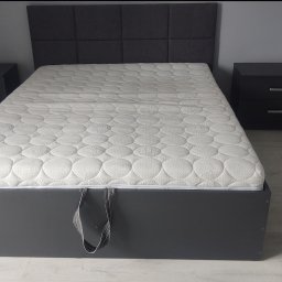 DZIAŁAK BOGDAN KARPENTER FIRMA WIELOBRANŻOWA - Szare łóżko z tapicerowanym zagłówkiem i materacem, z szafkami nocnymi po bokach. W tle ściana i podłoga w odcieniach szarości. Widoczne narzędzia i elementy montażowe.